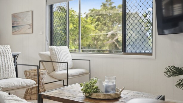 Una sala de estar acogedora con una mesa de centro de madera, sillas blancas acolchadas y vistas a través de una ventana con mosquitero.