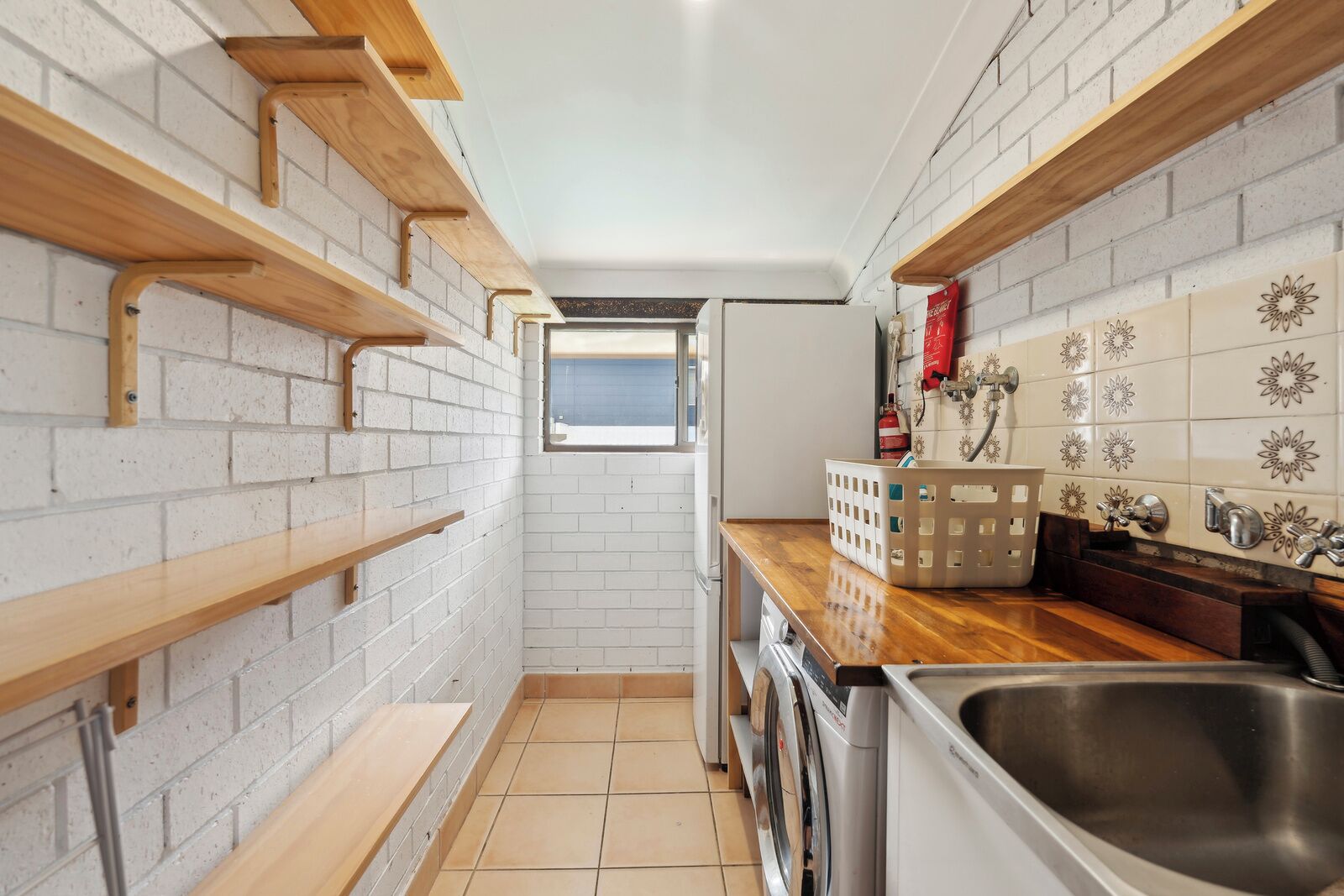 Een compacte keuken met witte bakstenen muren, houten planken, een raam, een wasmachine en een spoelbak.