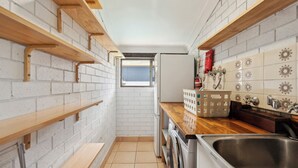 Una cocina compacta con paredes de ladrillo blanco, estantes de madera, una ventana, lavadora y un lavabo.