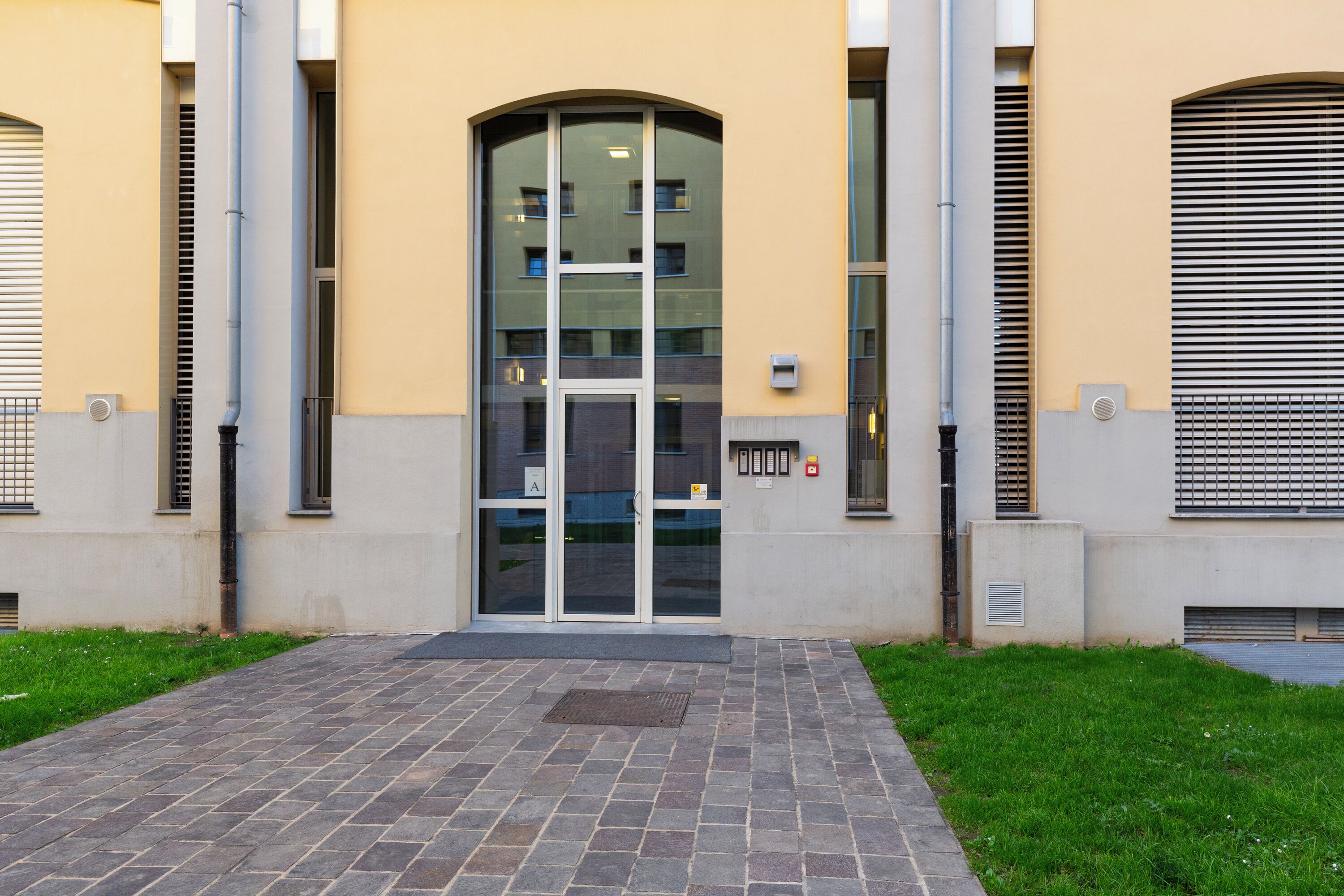 A building entrance with a glass door, a paved walkway, and a small grassy area.