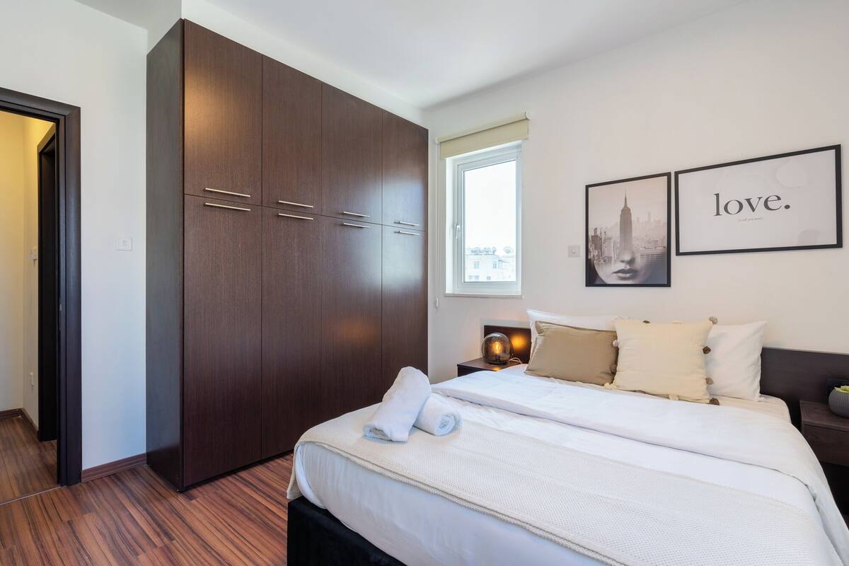 A bedroom with a large wardrobe, a bed with white bedding, a nightstand with a lamp, and two framed pictures on the wall.