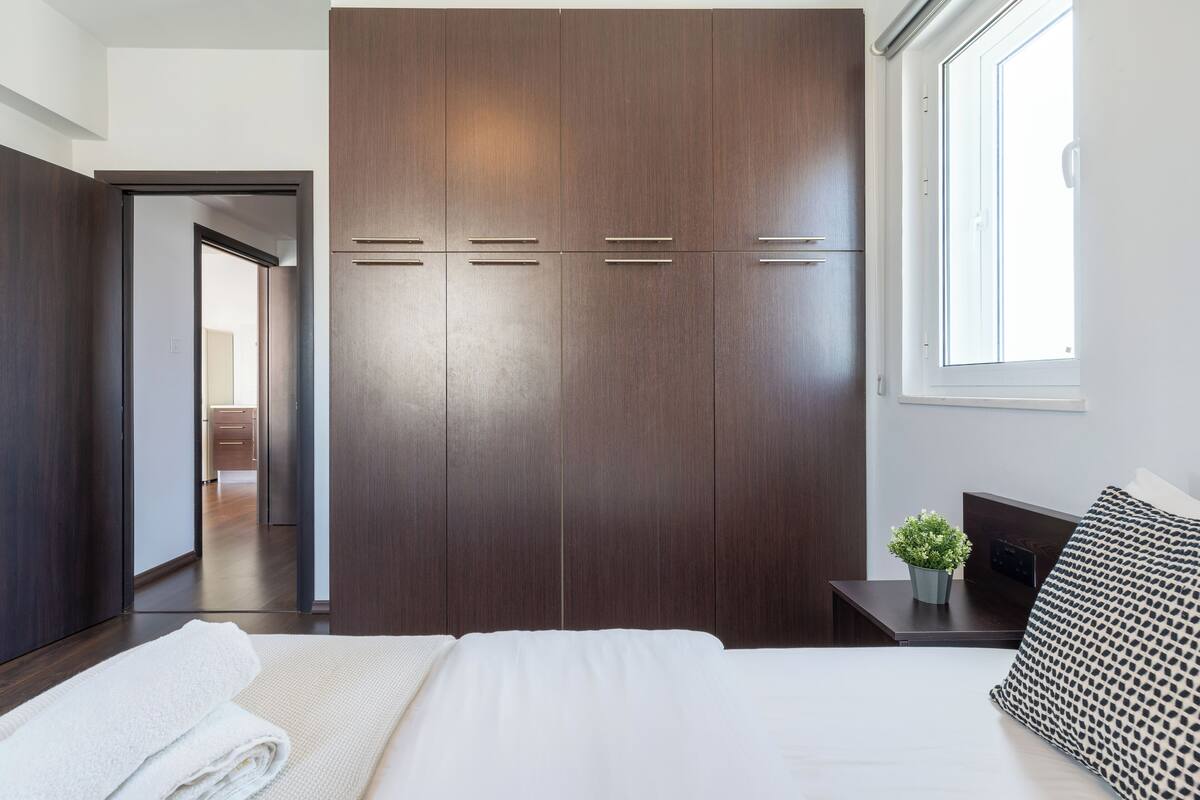 A bedroom with a large wardrobe, a bed with white bedding, a small bedside table with a plant, and a window with natural light.