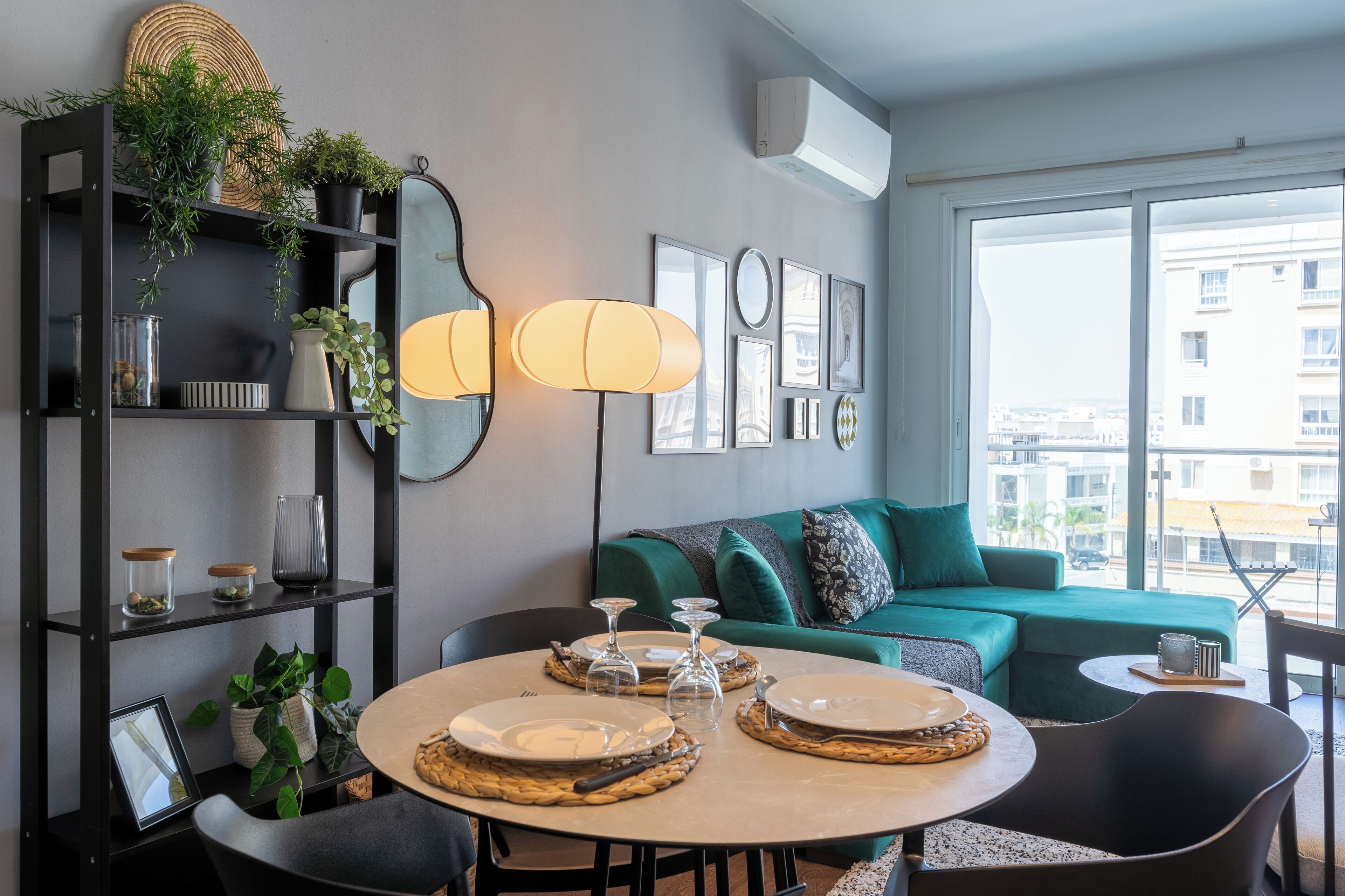 A modern living room with a round dining table set for four, a green sofa, and a large window offering a city view.