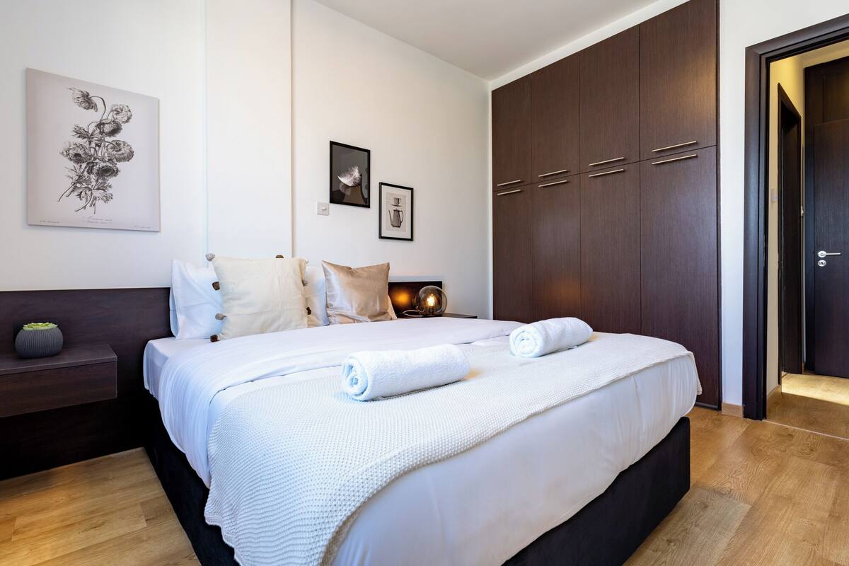 A neatly made bed with white linens, a dark wooden wardrobe, and a framed botanical print on the wall.