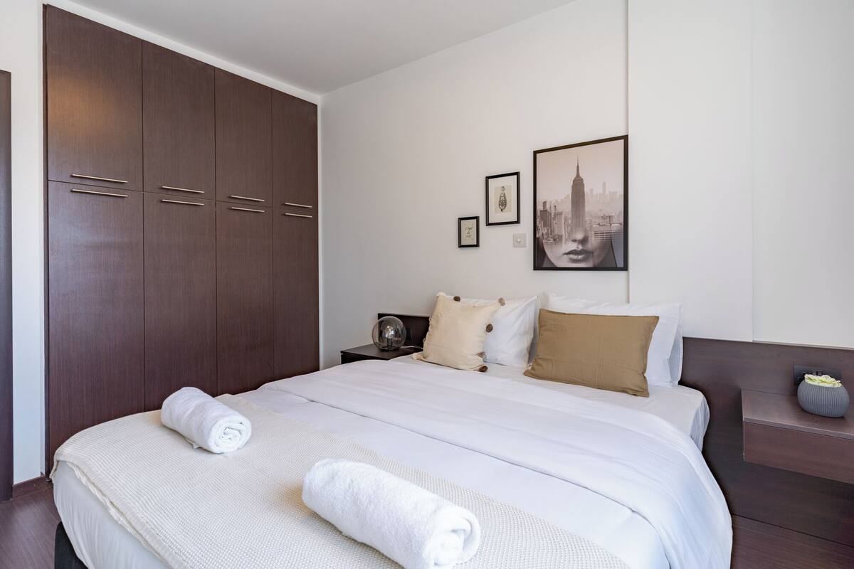 A hotel room with a large bed, a wardrobe, a nightstand, and a framed cityscape picture on the wall.