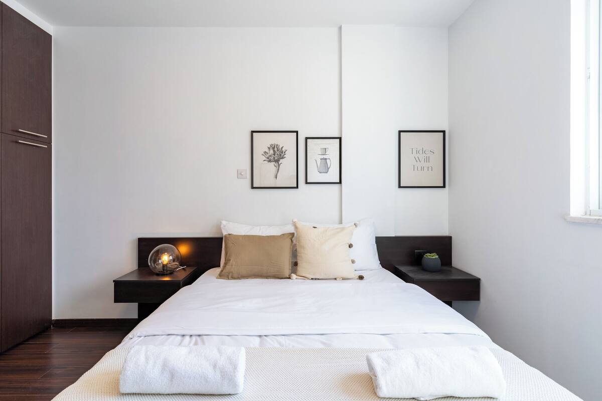 A modern bedroom with a large bed, two bedside tables, and a nightstand with a lamp. There are framed pictures on the wall and a window on the right side.