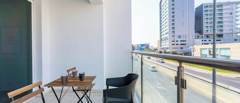 A balcony with a wooden table and chairs, overlooking a city street and tall buildings.