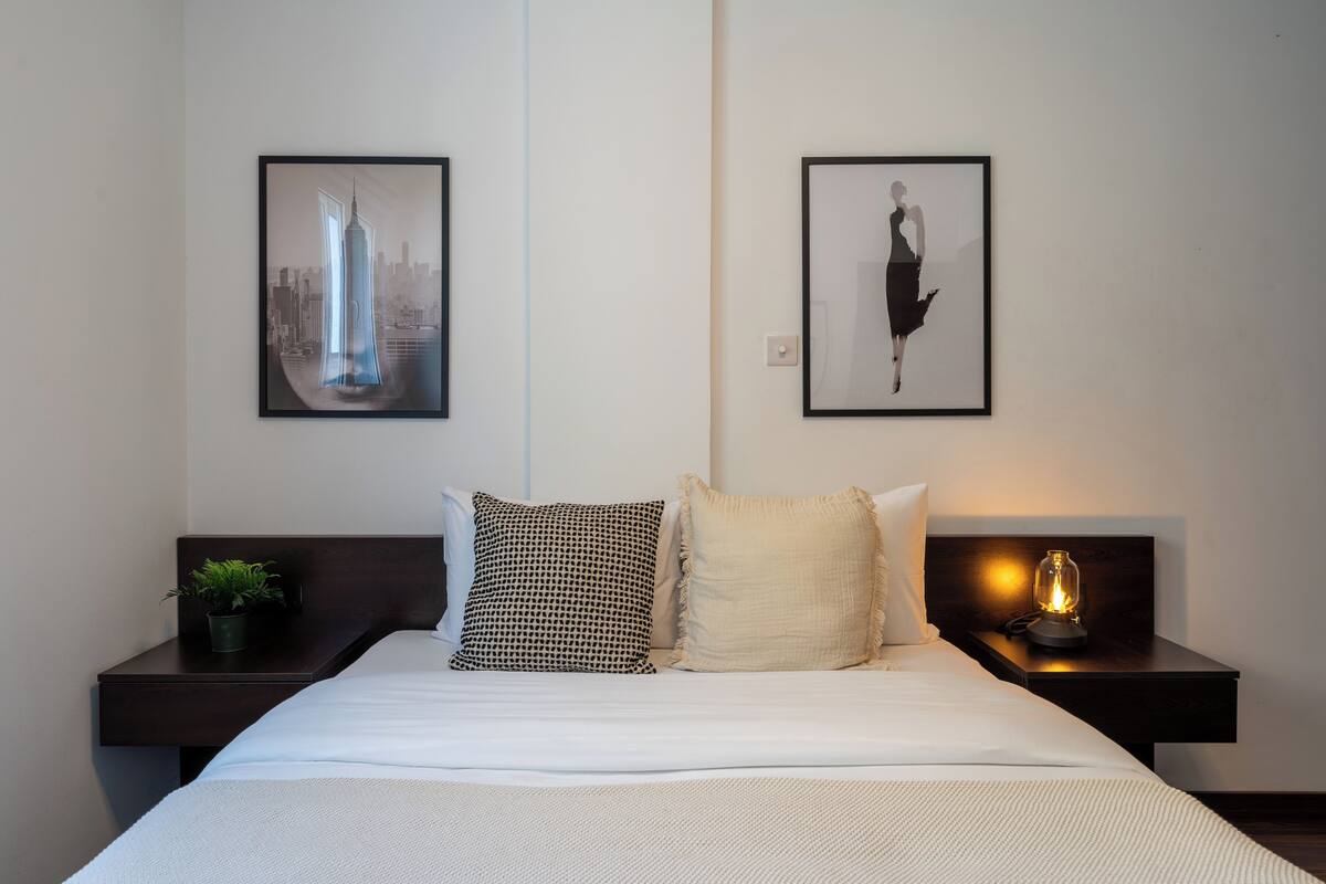 A neatly made bed with white bedding, two pillows, a small potted plant on the left side, and a nightstand with a lamp on the right.