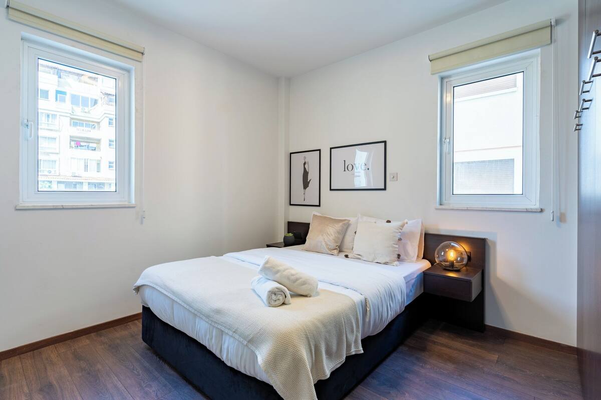 A bedroom with a bed, two bedside tables, a wall-mounted lamp, and two framed pictures.