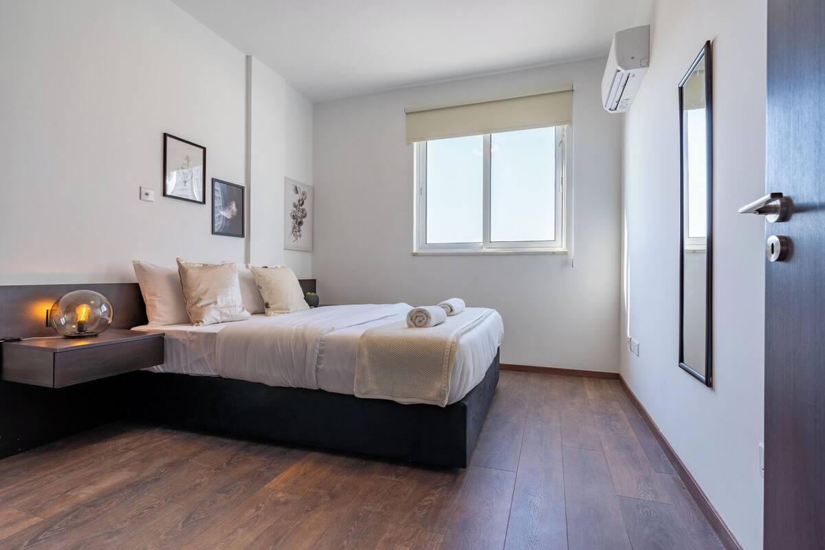 A bedroom with a bed, a nightstand, a wall-mounted air conditioner, and a window.