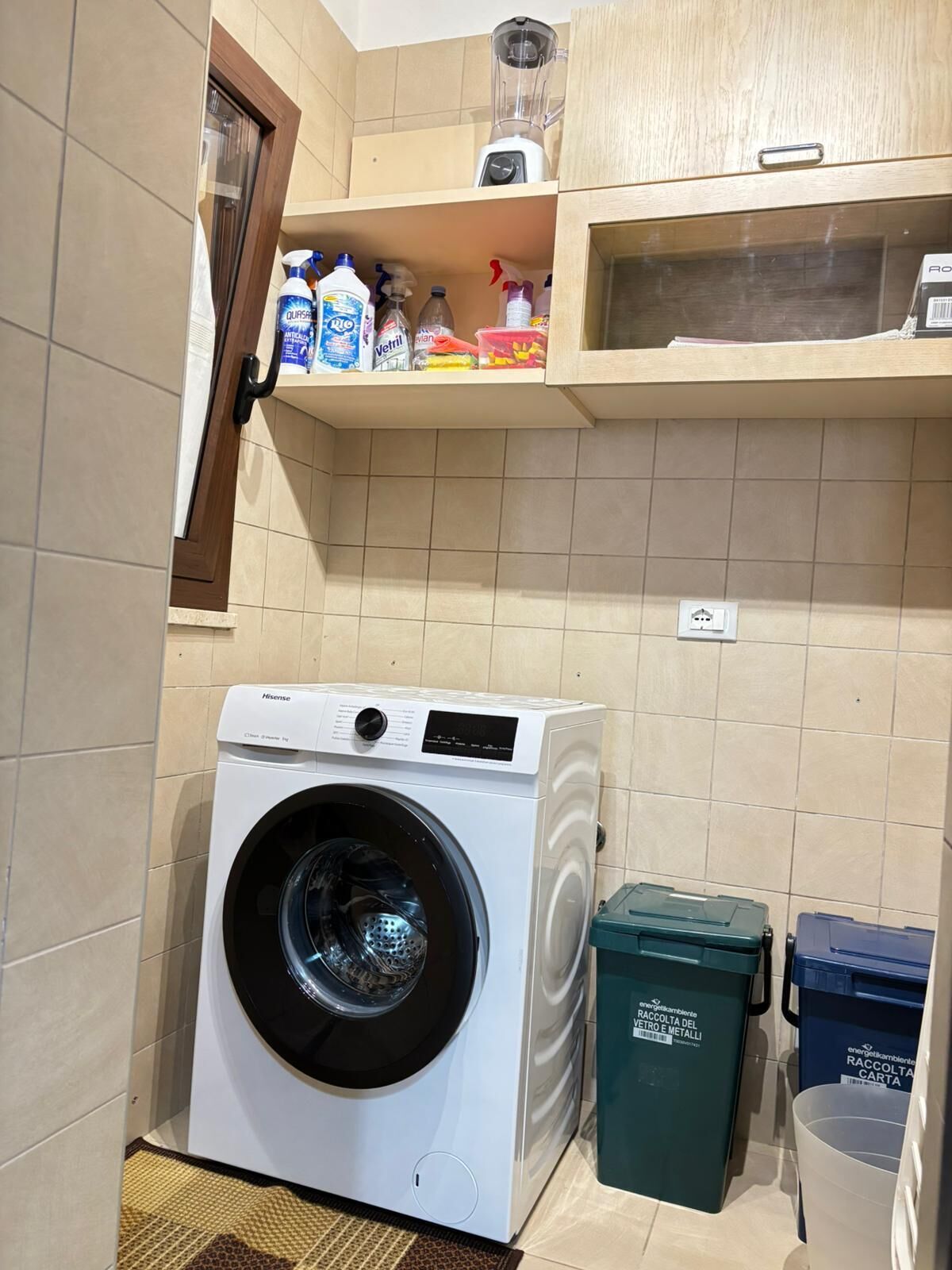 Ein kleiner Waschraum mit einer Waschmaschine mit Frontlader, einer Ablage für Reinigungsmittel und Recyclingbehältern.