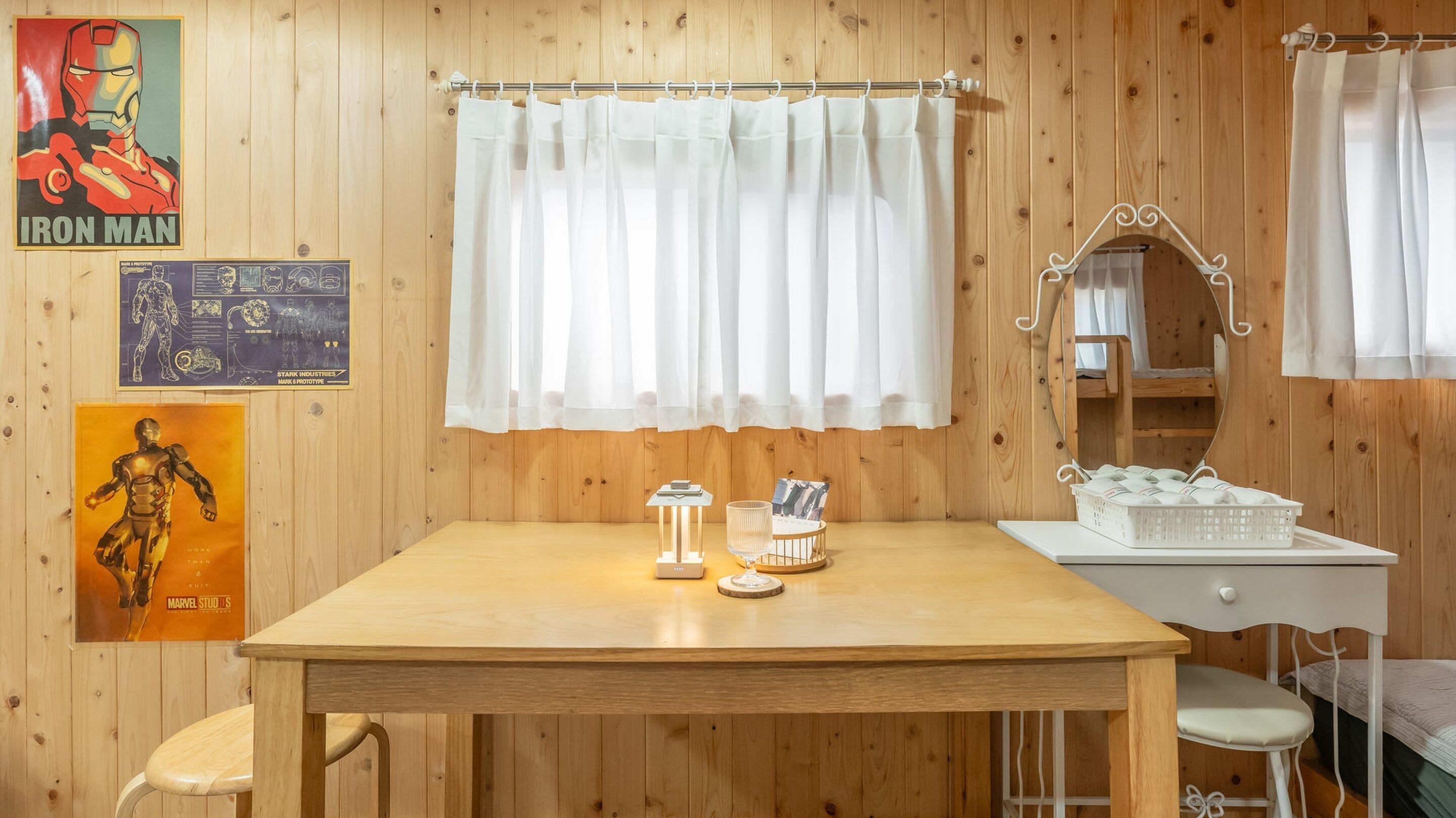 A wooden room with a table, a mirror, and a bed. There are posters on the wall and a curtain over the window.
