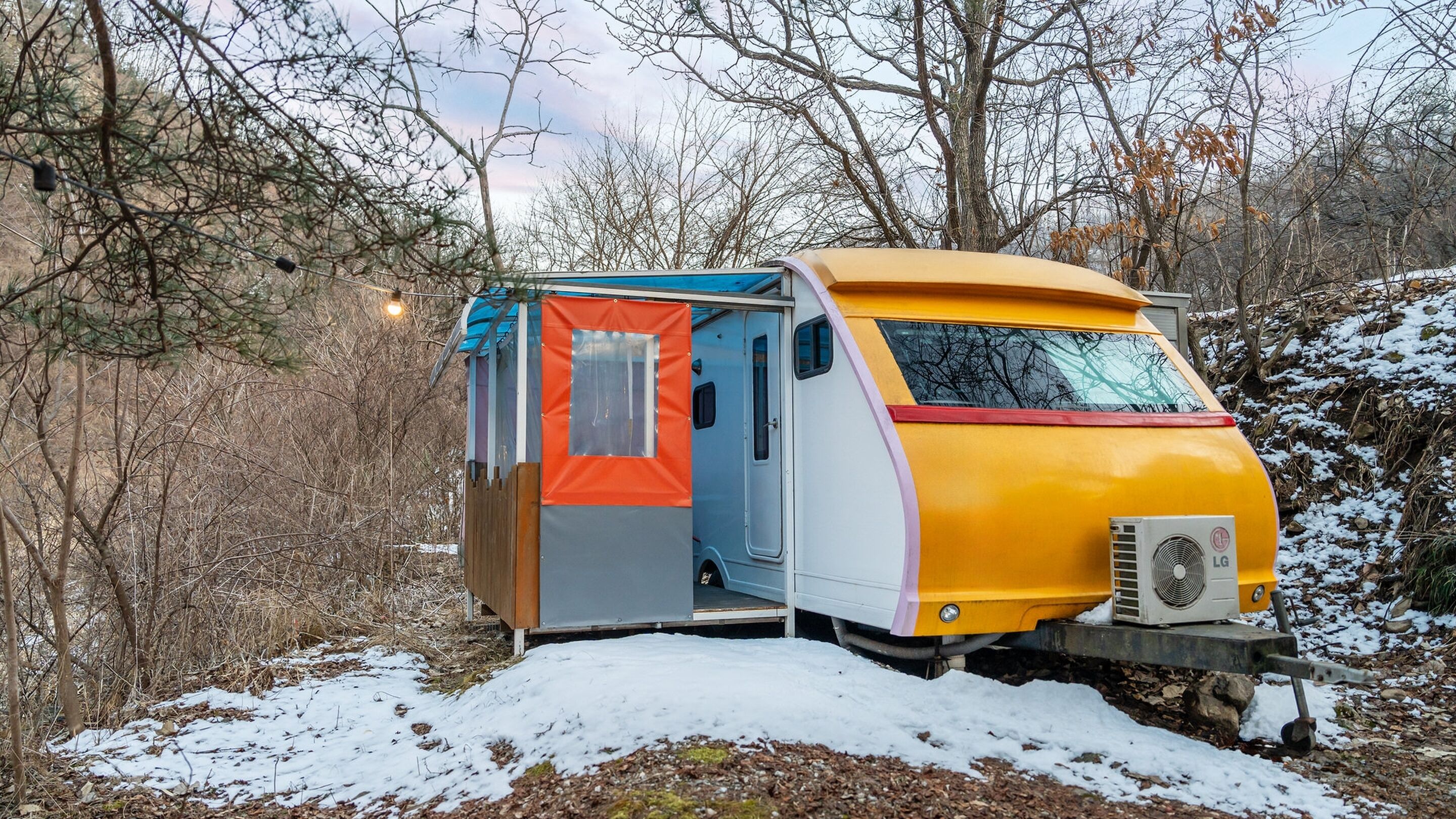 Uma caravana pequena e fechada com porta deslizante, estacionada em uma área florestal coberta de neve.