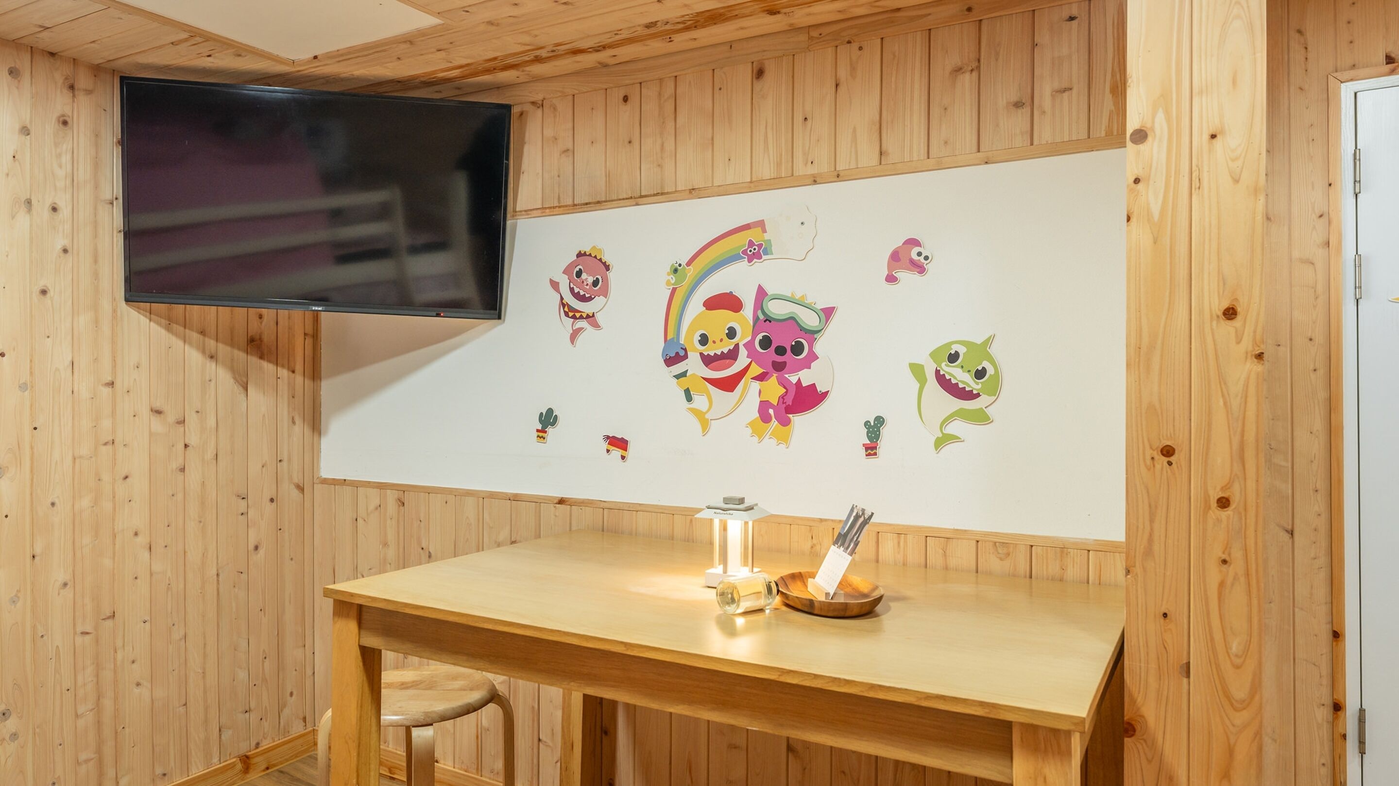 A wooden room with a flat-screen TV mounted on the wall, a wooden table with a glass bottle and a bowl, and playful wall decals.