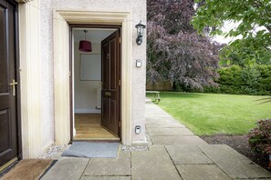 A house entrance with a wooden door, a doormat, and a pathway leading to a garden.