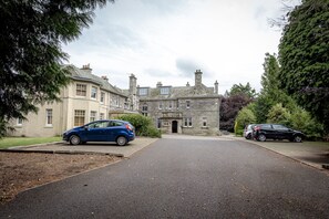 A stone building with a central entrance, two parked cars, and surrounding greenery.