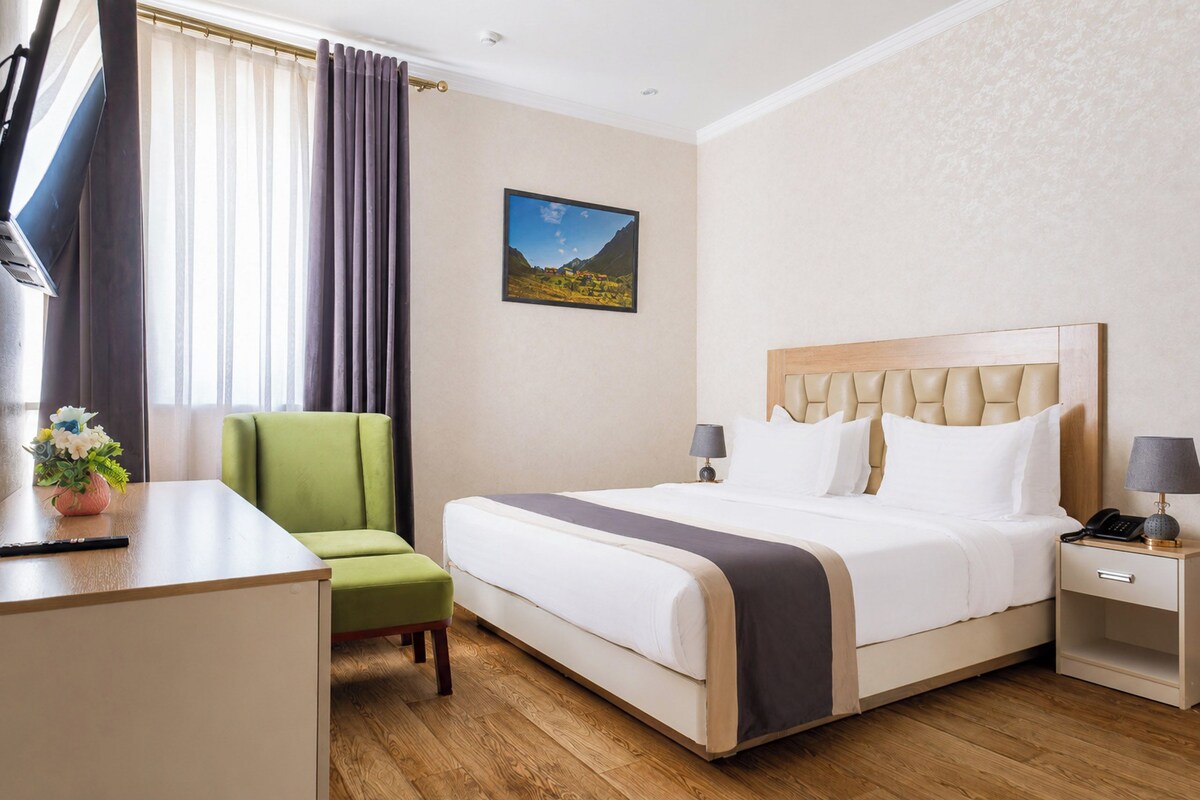 A hotel room with a bed, a desk, a chair, a nightstand, a lamp, and a picture on the wall.