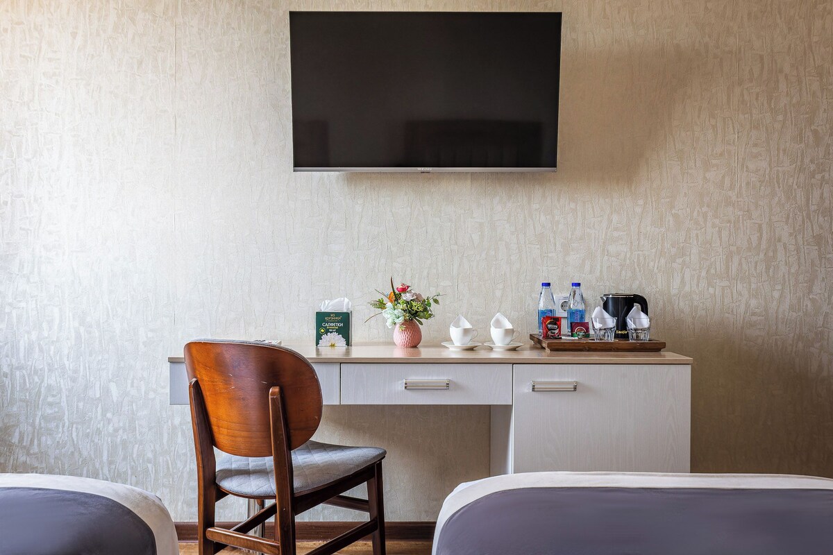 A hotel room with a bed, a desk with a chair, a television mounted on the wall, and a small table with a vase of flowers and various items.