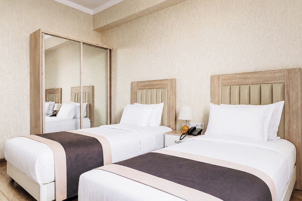 A hotel room with two beds, a large mirror, a bedside table with a lamp, and a telephone.