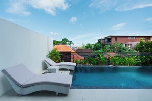 A swimming pool with lounge chairs, a white wall, and a building in the background.