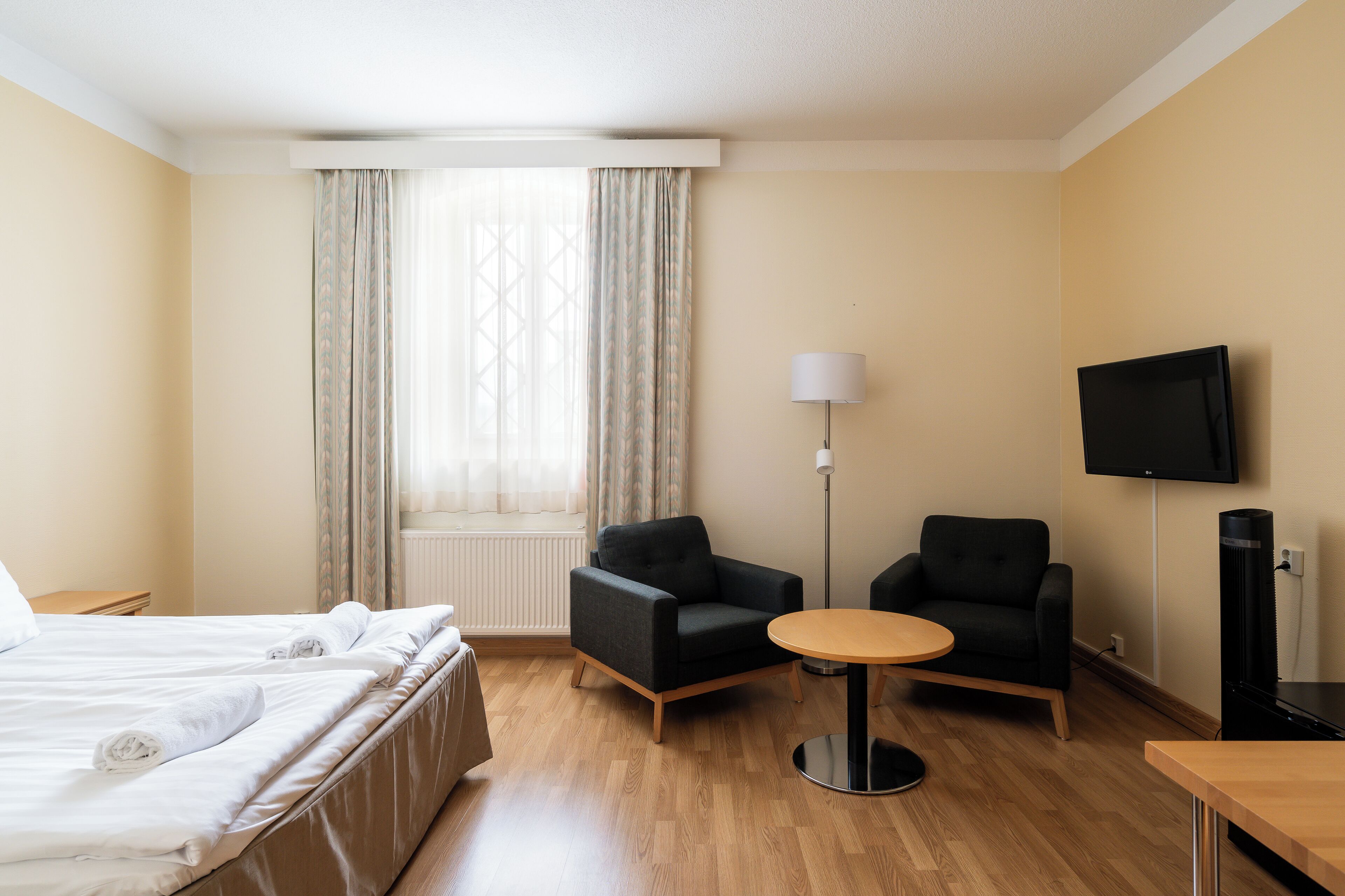 Una habitación de hotel con una cama, dos sillones, una mesita redonda, una lámpara y un televisor.
