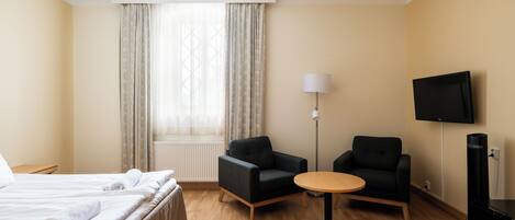 Una habitación de hotel con una cama, dos sillones, una mesita redonda, una lámpara y un televisor.