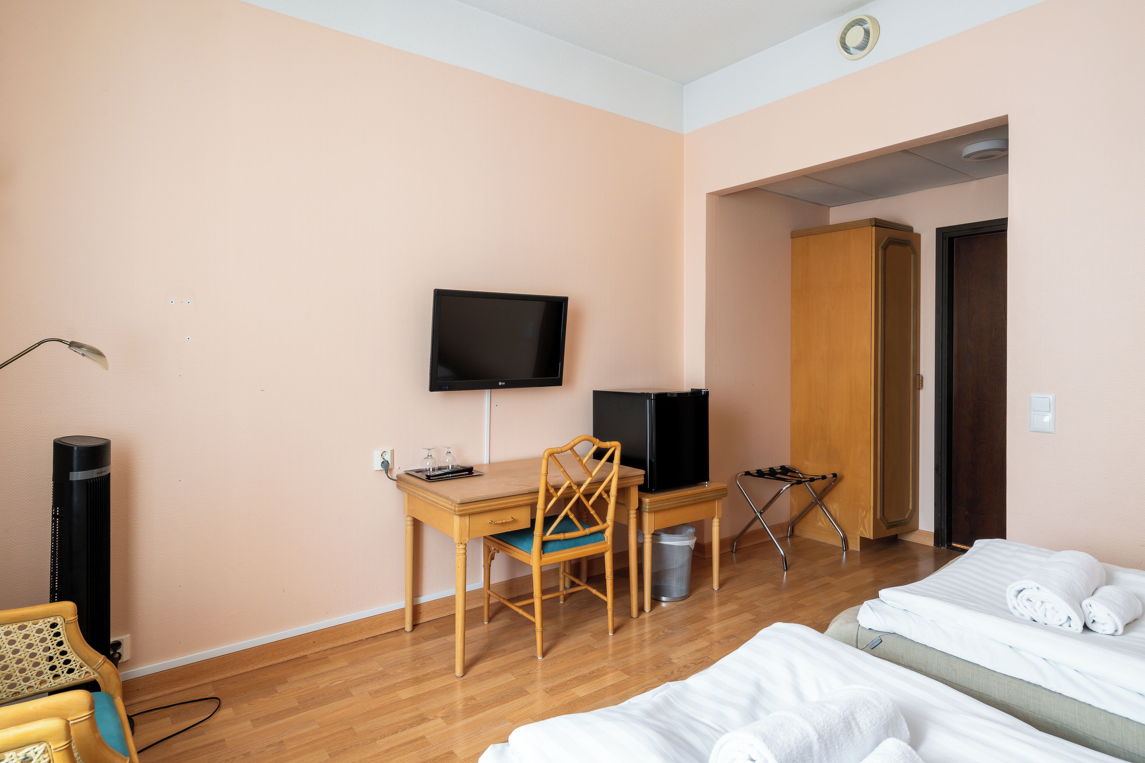 A hotel room with a wooden desk, a television, a chair, a bed with towels, and a trash bin.