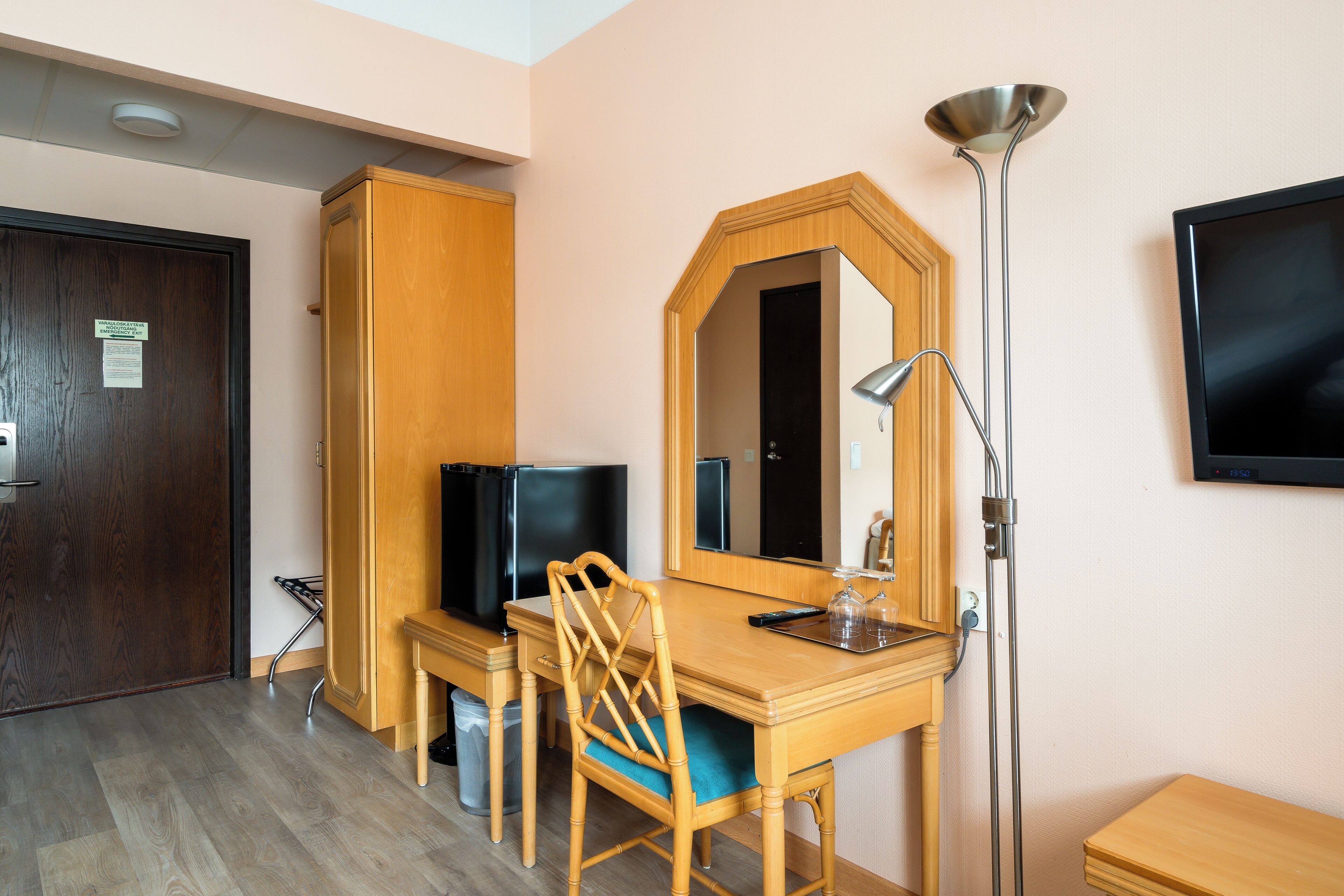 A hotel room with a wooden desk, a chair, a television, a mirror, a lamp, and a trash bin.