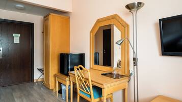 A hotel room with a wooden desk, a chair, a television, a mirror, a lamp, and a trash bin.