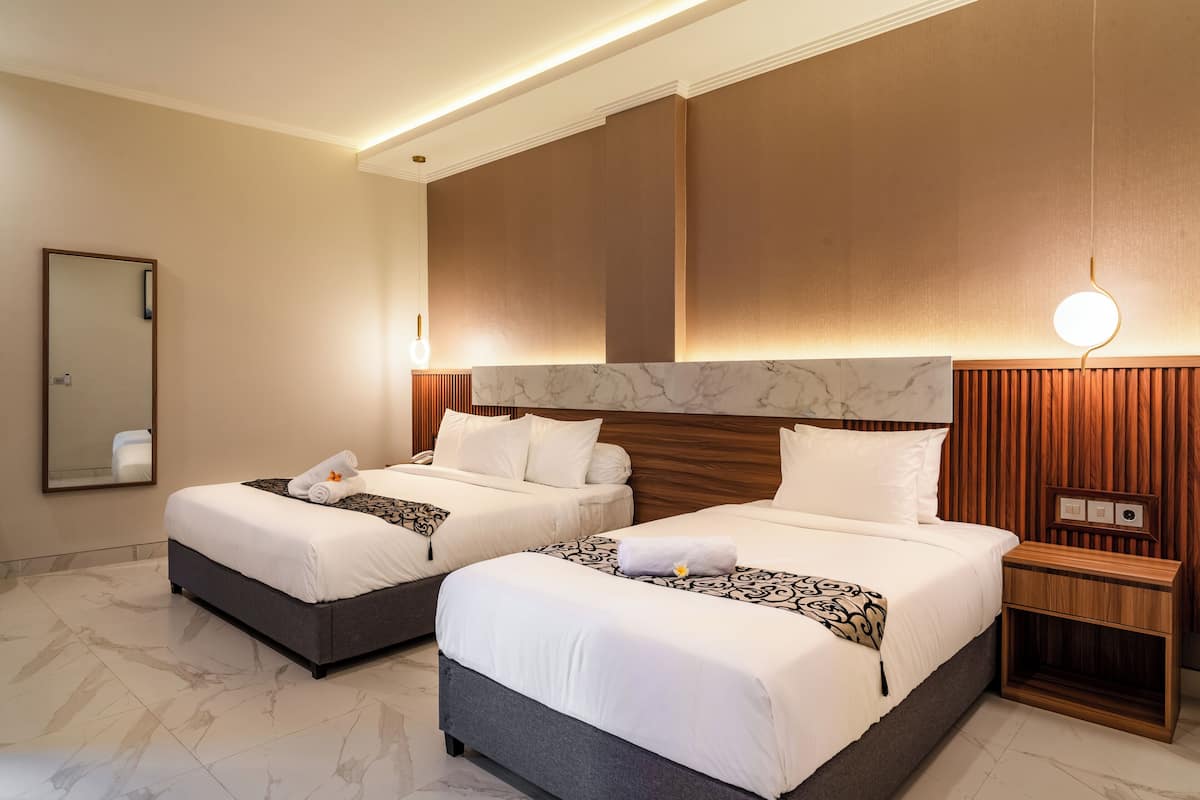 A hotel room with two beds, marble flooring, wooden headboard, and a nightstand with a lamp.