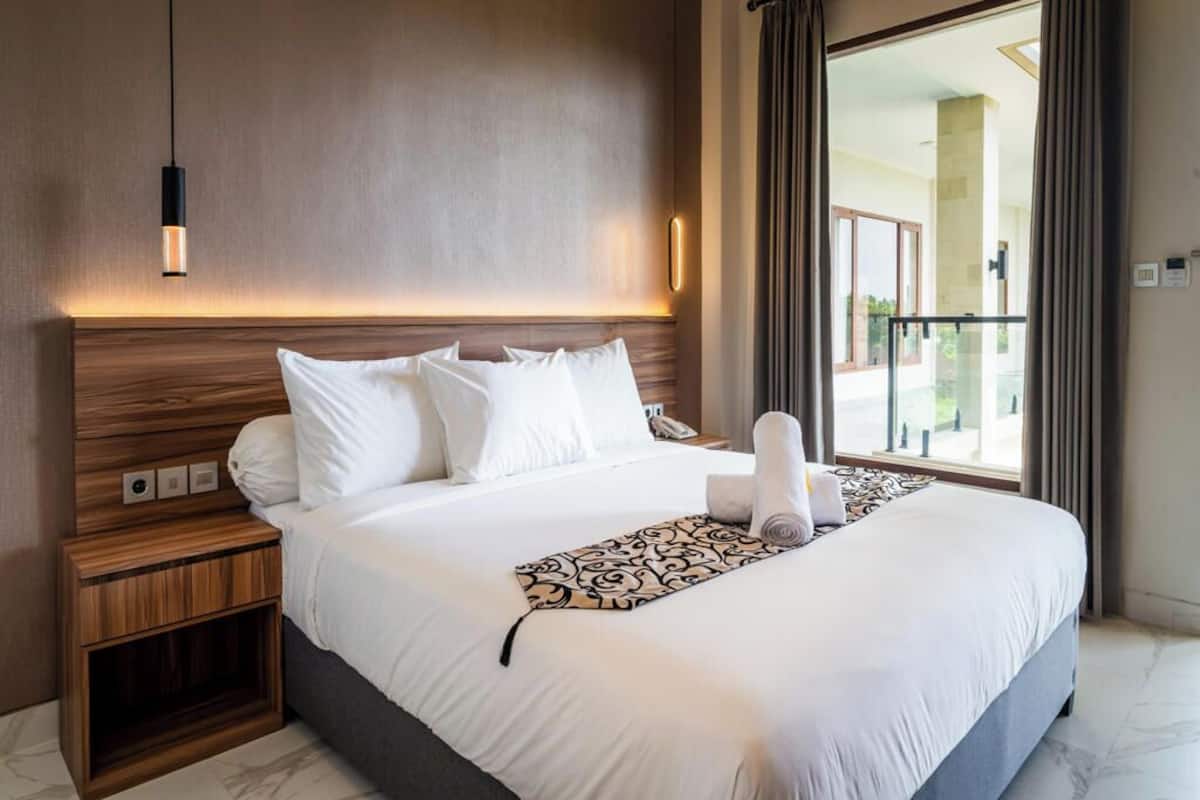 A hotel room with a large bed, a wooden nightstand, a modern pendant light, and a view of the outdoors through a large window.