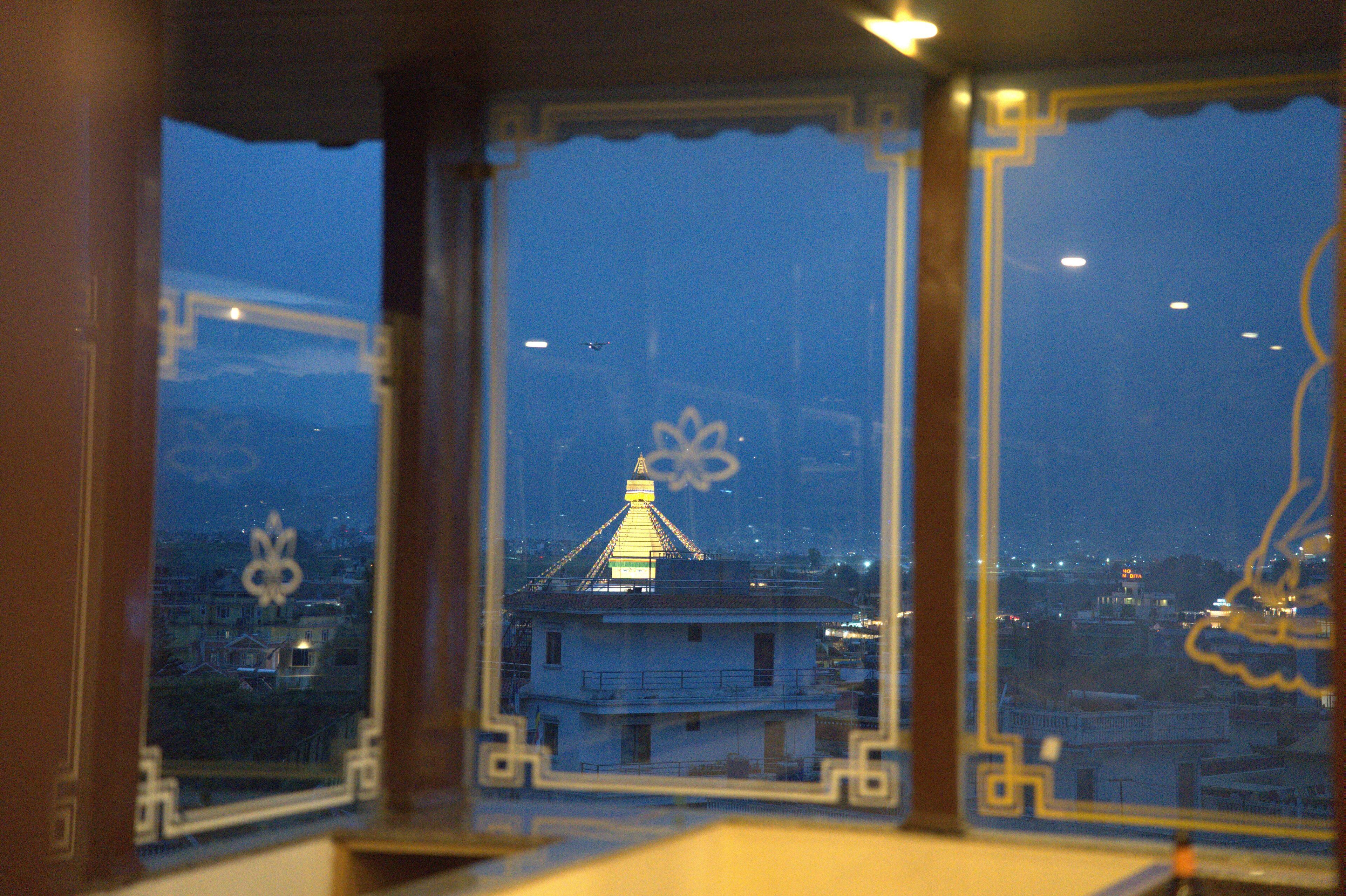 Vista a través de una ventana con un marco decorativo, que muestra un edificio con una torre iluminada y un paisaje urbano al fondo.