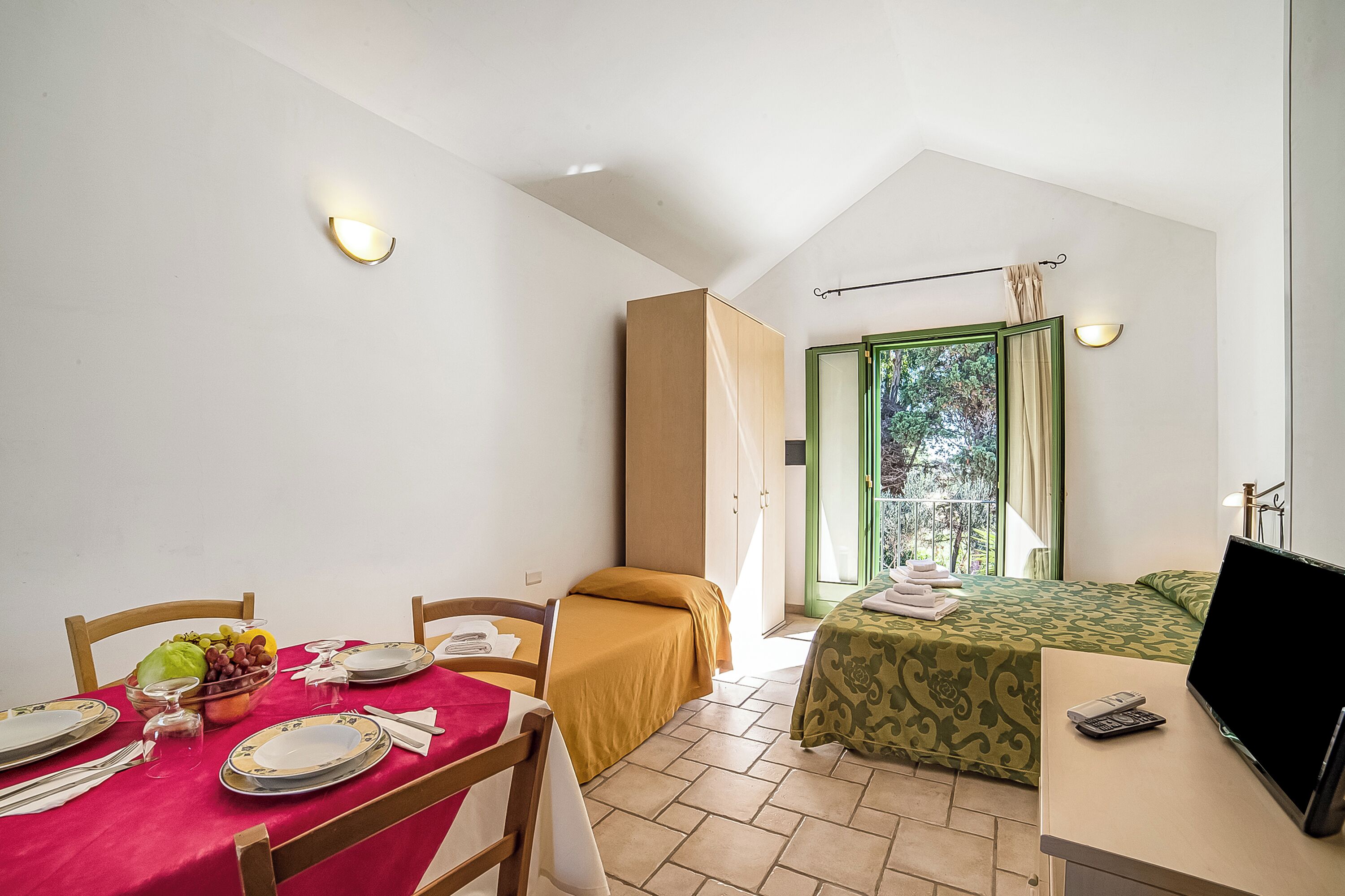 A room with a dining table set for two, a bed with a green bedspread, a TV on a desk, and a window with a view of greenery.