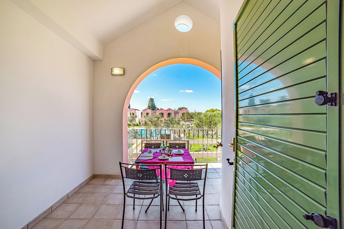 Un balcon avec une table dressée pour quatre personnes, une porte à volets verts et une vue sur les bâtiments et la verdure.