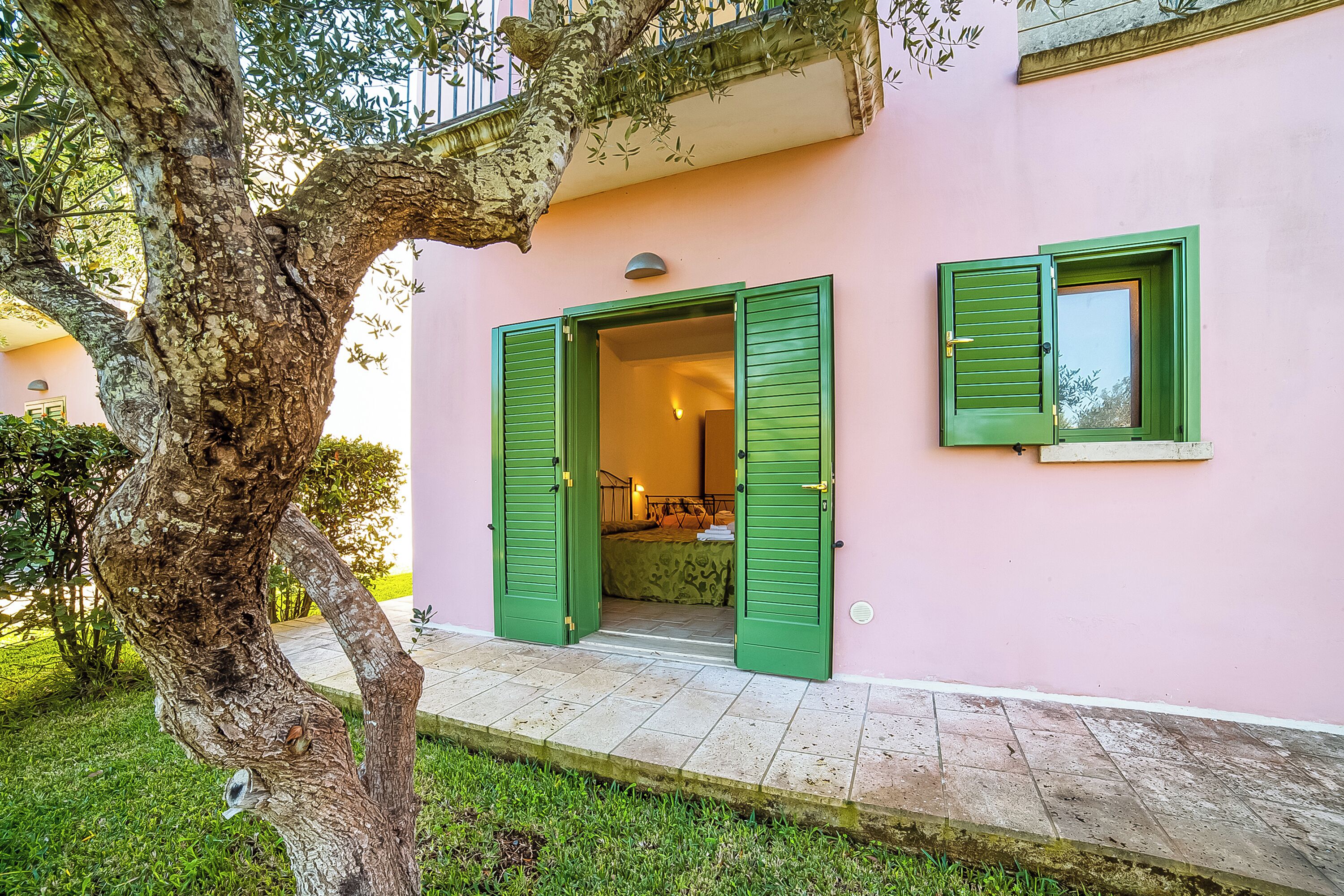 A pink building with green shutters, a visible room with a bed, and a tree in the foreground.