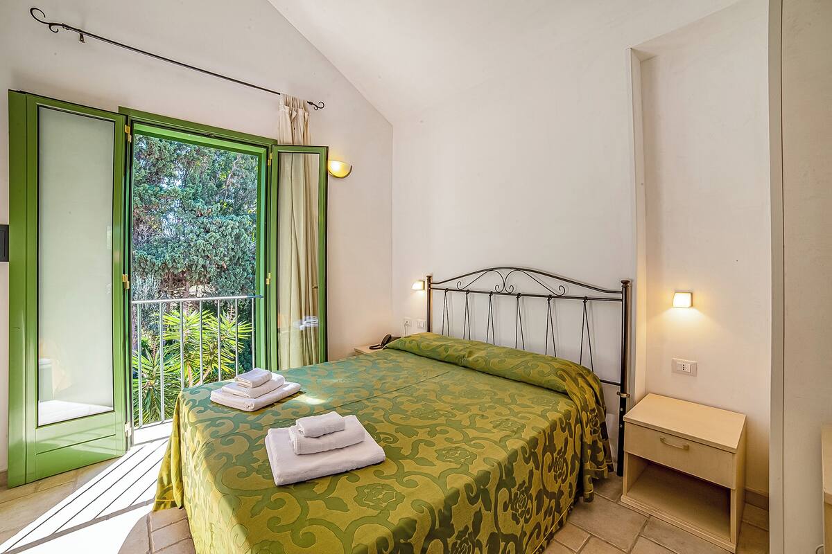 Une chambre à coucher avec une couvre-lit à motifs verts, une table de chevet et une vue sur la verdure grâce à une porte coulissante.
