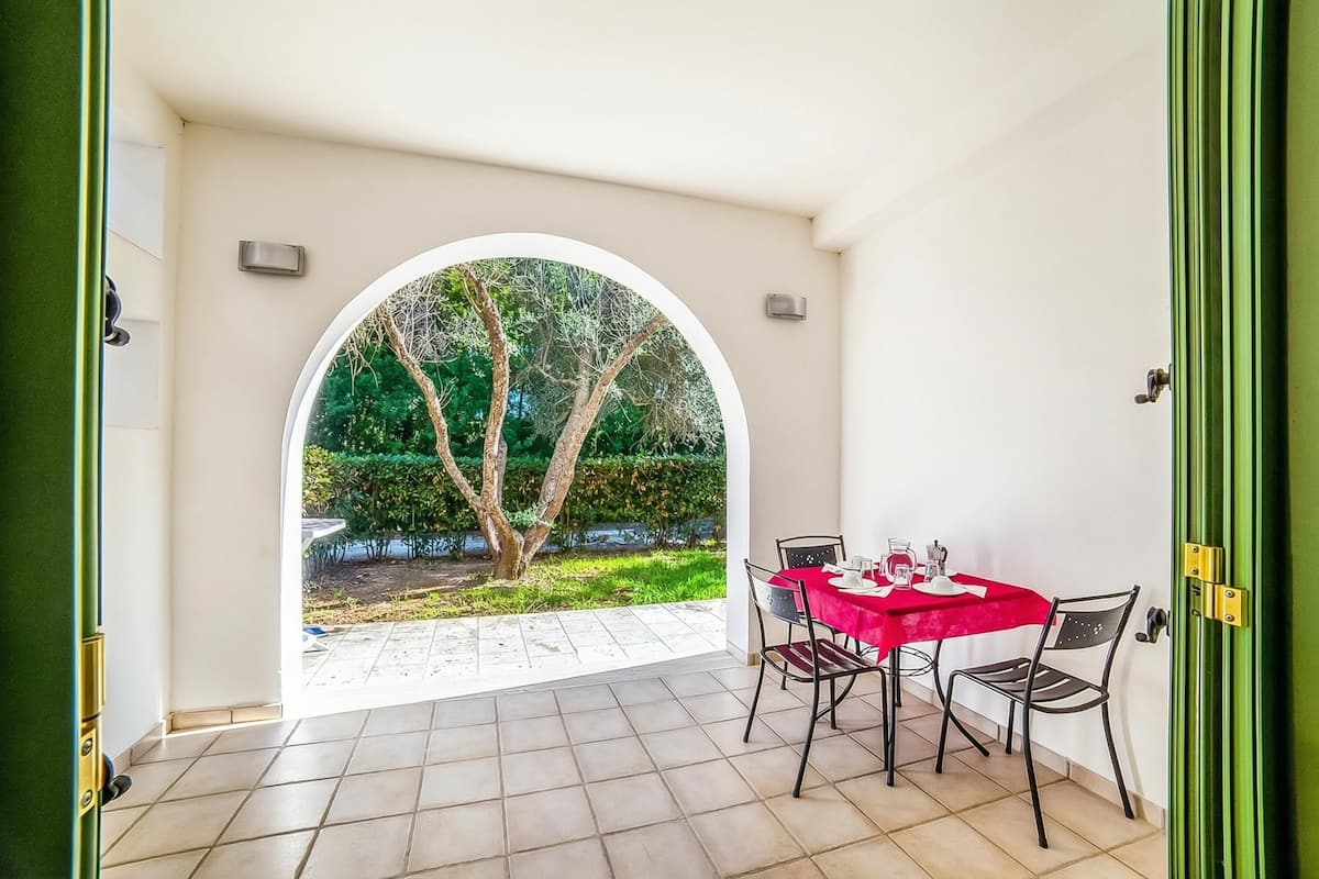 Une pièce avec un sol carrelé, une table recouverte d’une nappe rouge, deux chaises et, vue à travers une porte en arc de cercle, un jardin avec des arbres.
