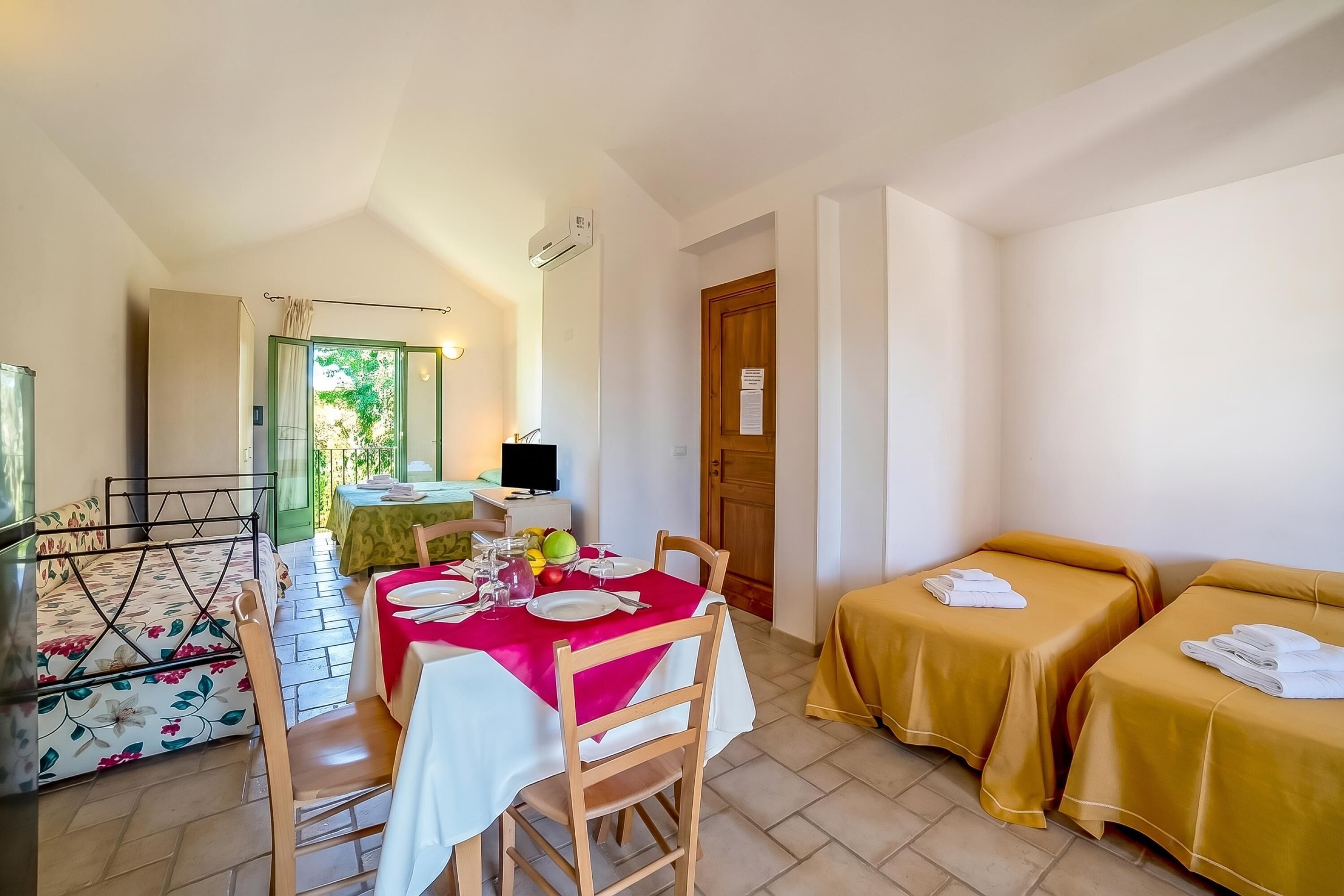 Una habitación con una mesa de comedor lista para cuatro personas, dos camas individuales, un televisor y una puerta de madera.
