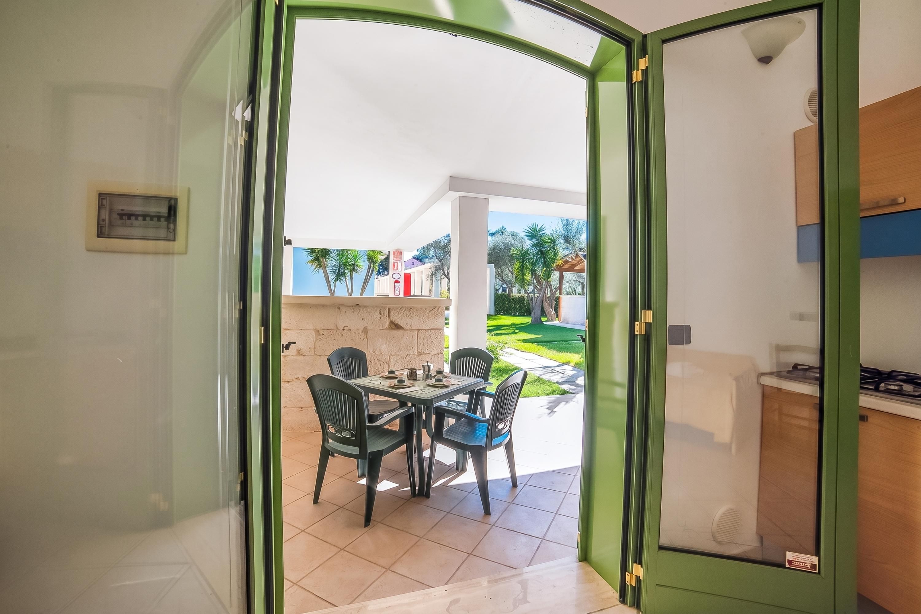 Uma porta aberta dá para um pátio com mesa e cadeiras, piso de azulejos e vista para um jardim com árvores e um edifício ao fundo.