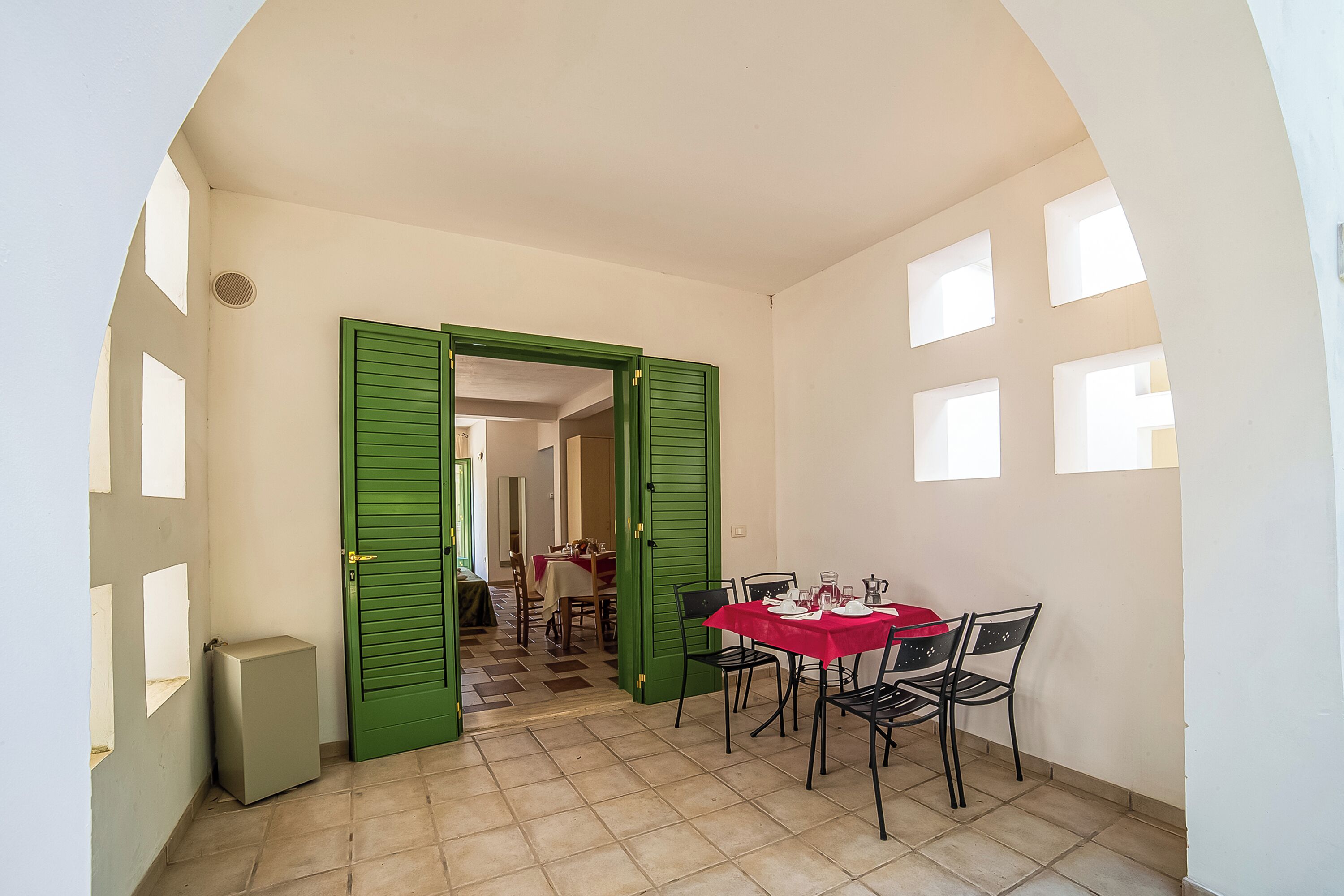 Un coin repas avec une table dressée pour quatre personnes, des volets verts sur la porte et un sol carrelé.