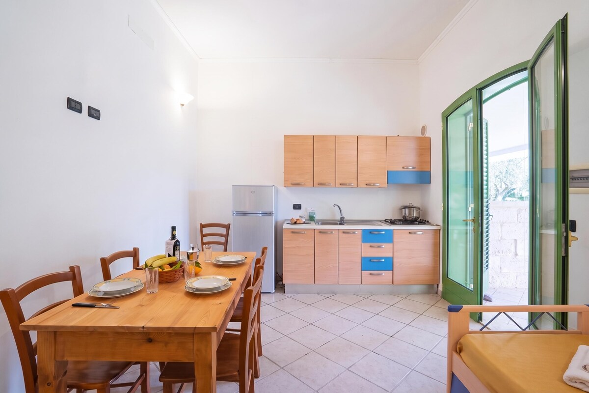 A compact kitchen with wooden cabinets, a dining table set for four, and a small refrigerator.