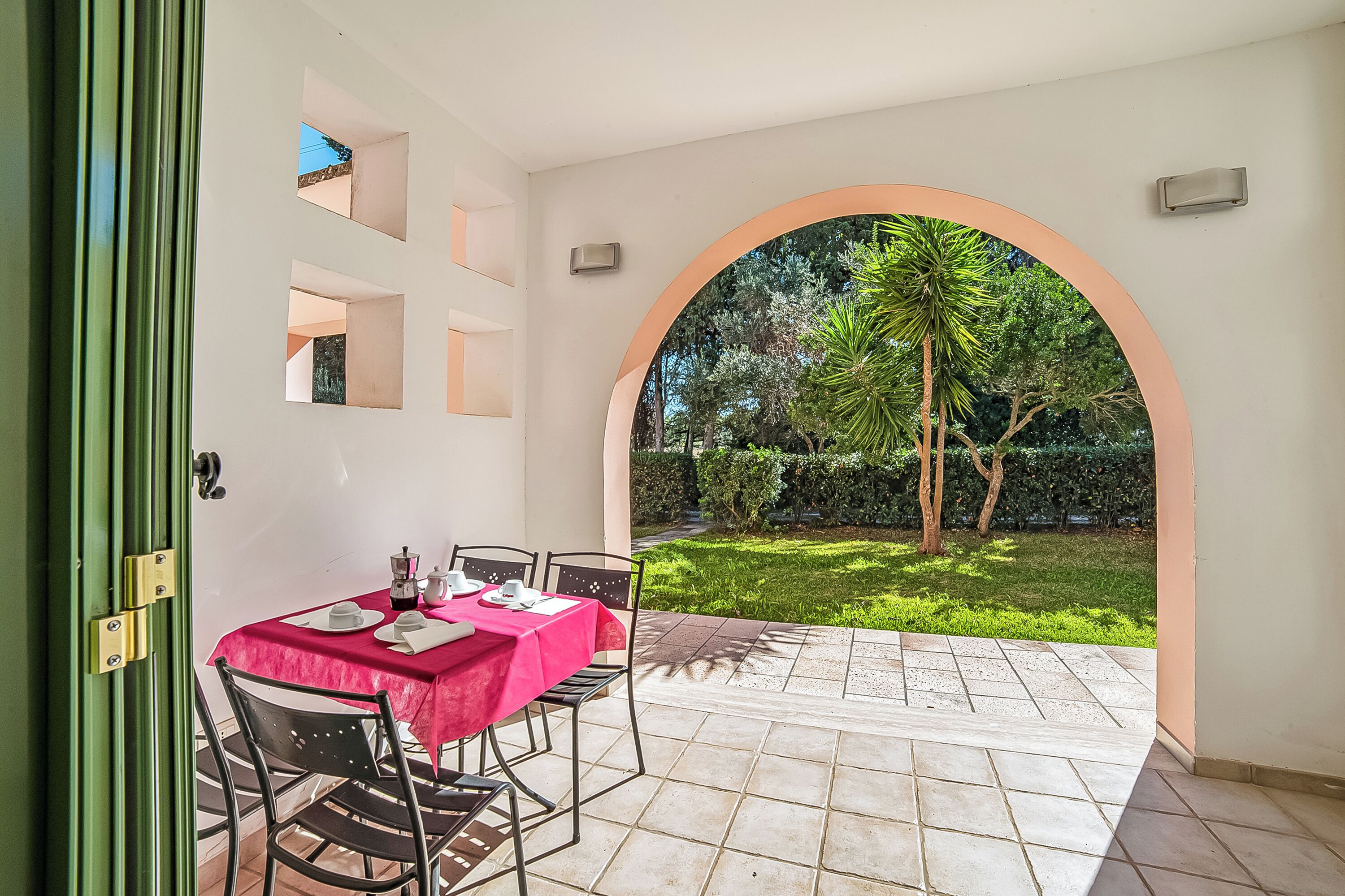 An outdoor patio with a table set for two, a green door, and a view of a garden through an archway.