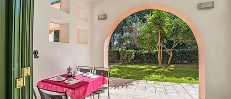 An outdoor patio with a table set for two, a green door, and a view of a garden through an archway.