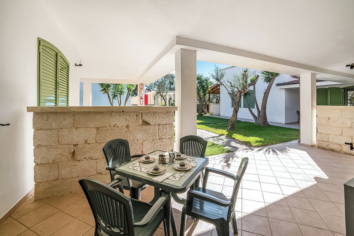 Une terrasse avec une table dressée pour quatre personnes, entourée de chaises, et une vue sur un jardin et des bâtiments en arrière-plan.