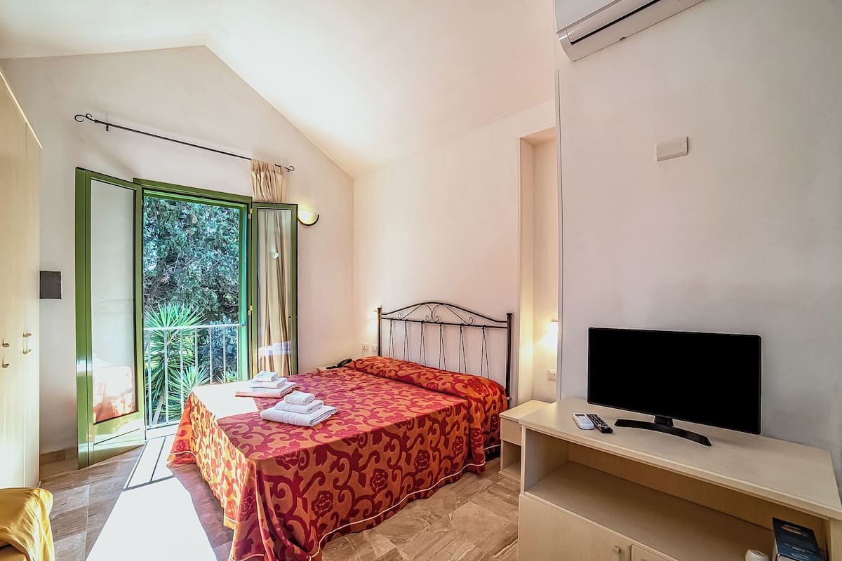 Une chambre à coucher comprenant un lit, une télévision, un bureau et un balcon donnant sur un espace verdoyant.
