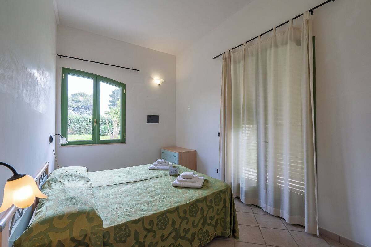 A bedroom with a bed, a nightstand, a lamp, a window with curtains, and a view of greenery outside.