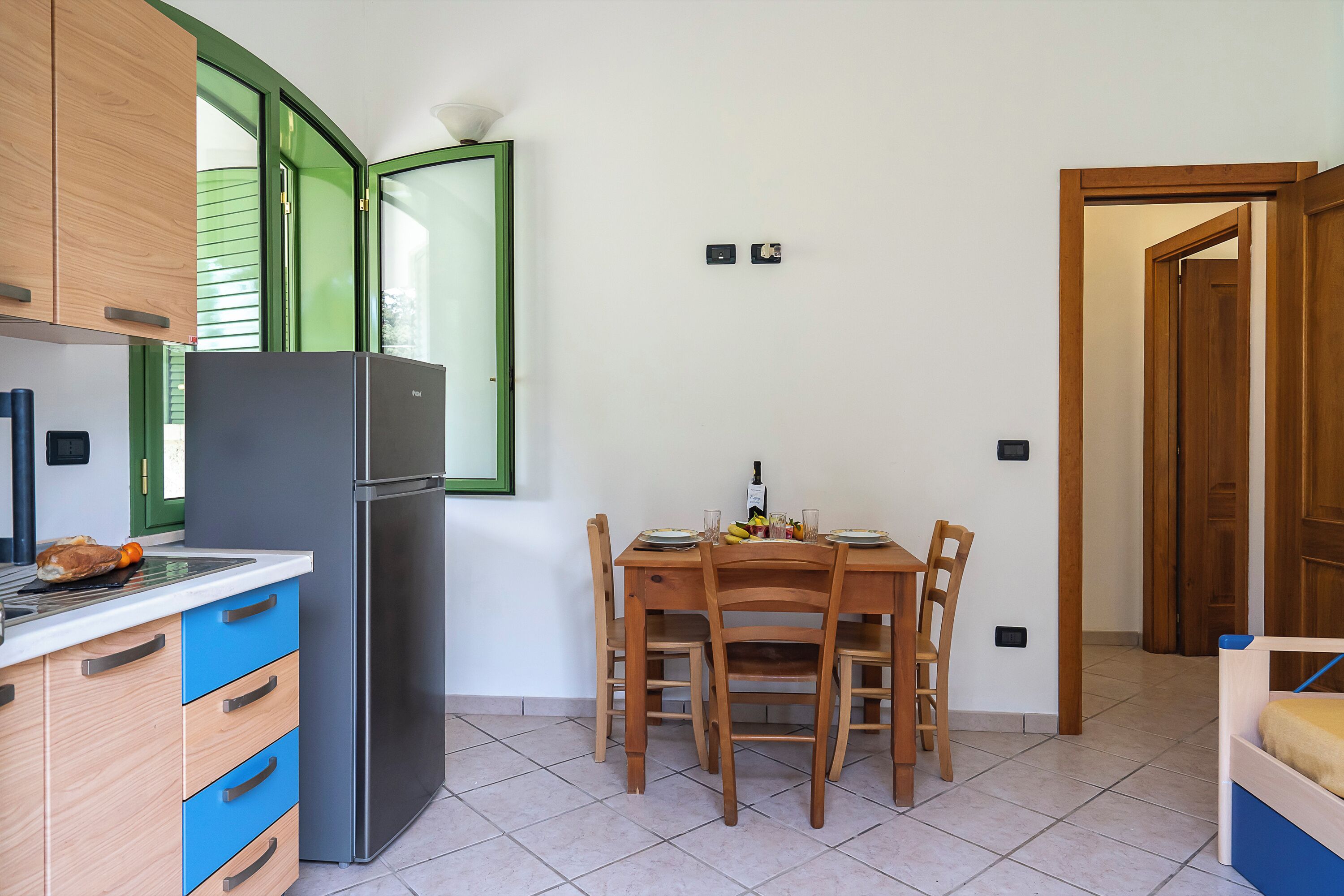A compact kitchen with a refrigerator, wooden cabinets, a dining table with chairs, and a window with green shutters.