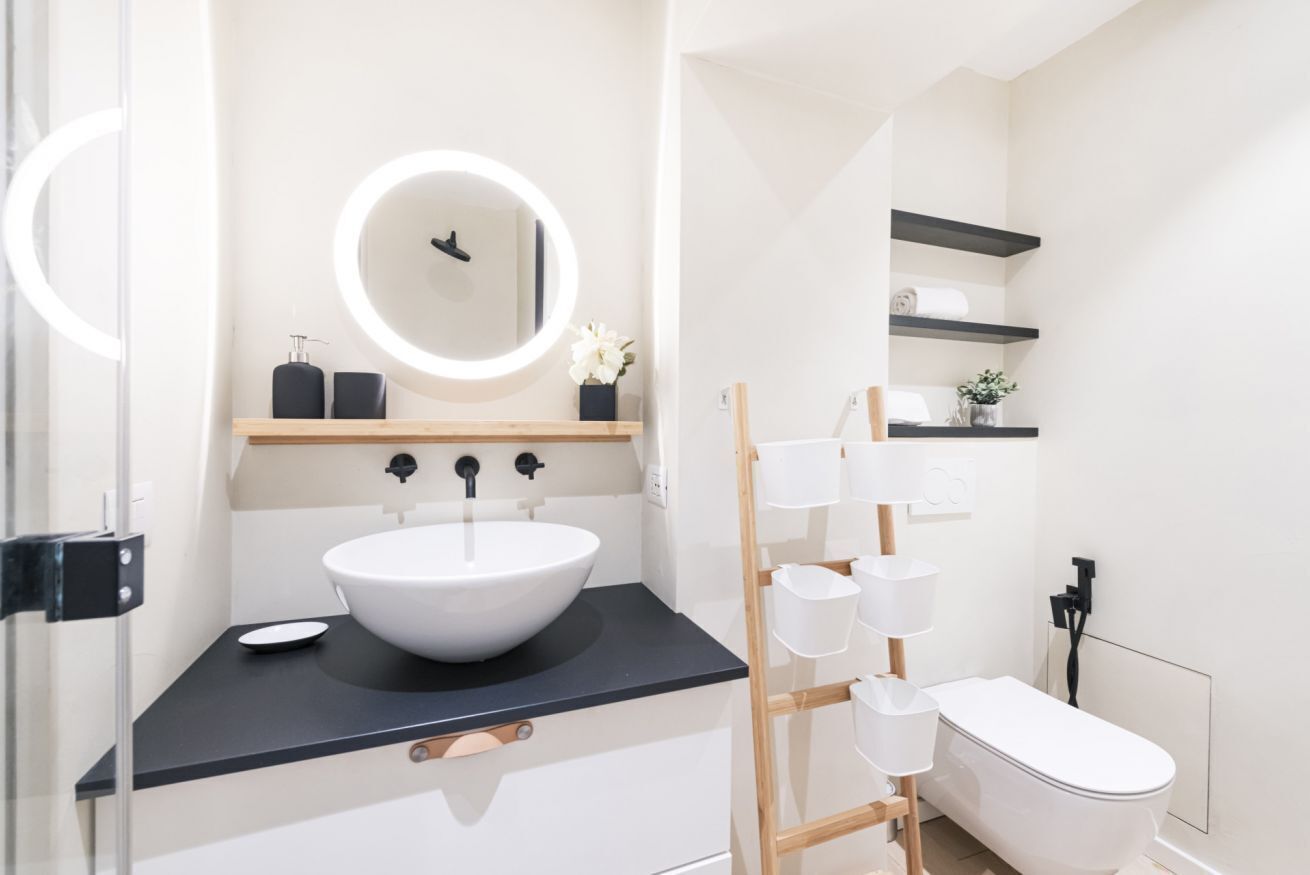 A modern bathroom with a white sink, black countertop, and a round mirror. There are shelves above the sink and a toilet with a wooden frame.
