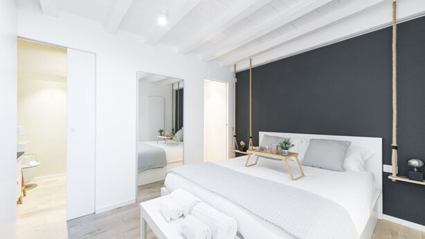 A modern bedroom with a large bed, bedside tables, a bench with towels, and a dark accent wall. - Easylife - Comfort and elegance in Porta Venezia (Milano)
