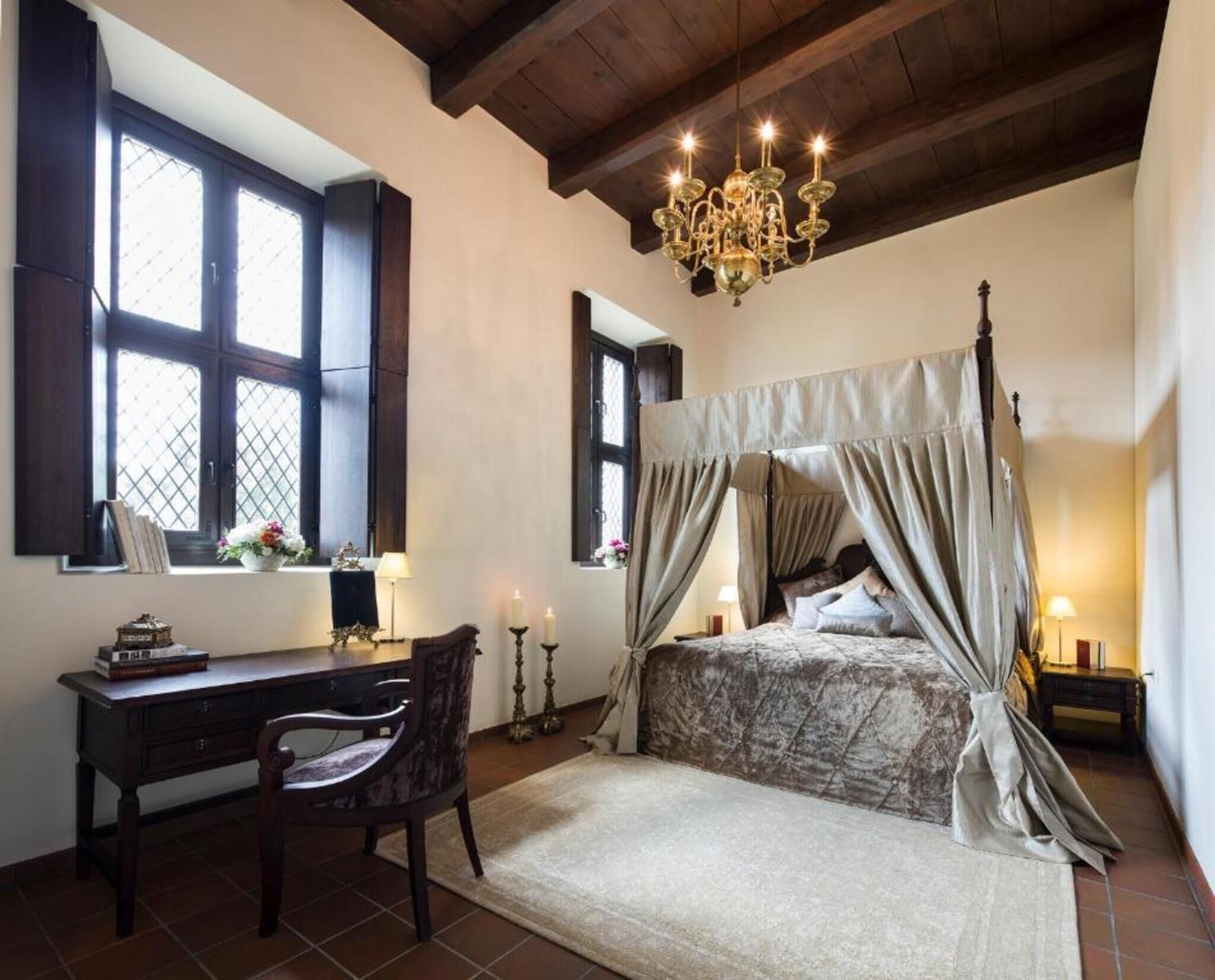 A bedroom with a four-poster bed, a desk with a chair, a chandelier, and a window with shutters.